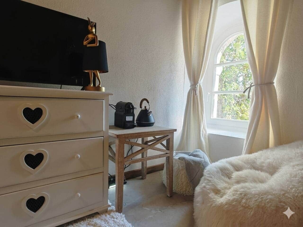 A room with a dresser with heart-shaped cutouts, a table with a kettle, and a fluffy white bed.