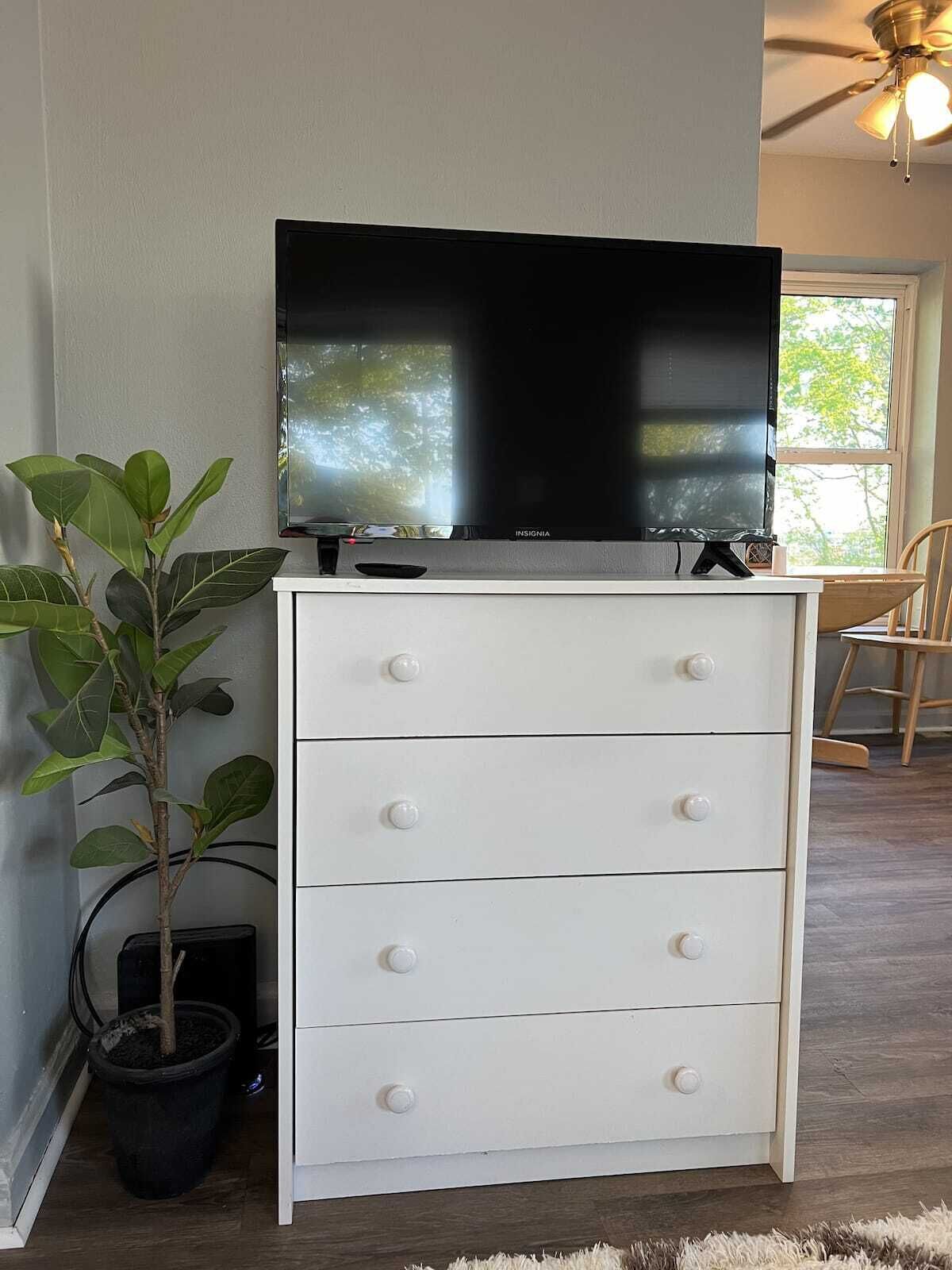 Une commode blanche avec des tiroirs, un téléviseur à écran plat posé dessus, une plante en pot et un ventilateur de plafond.