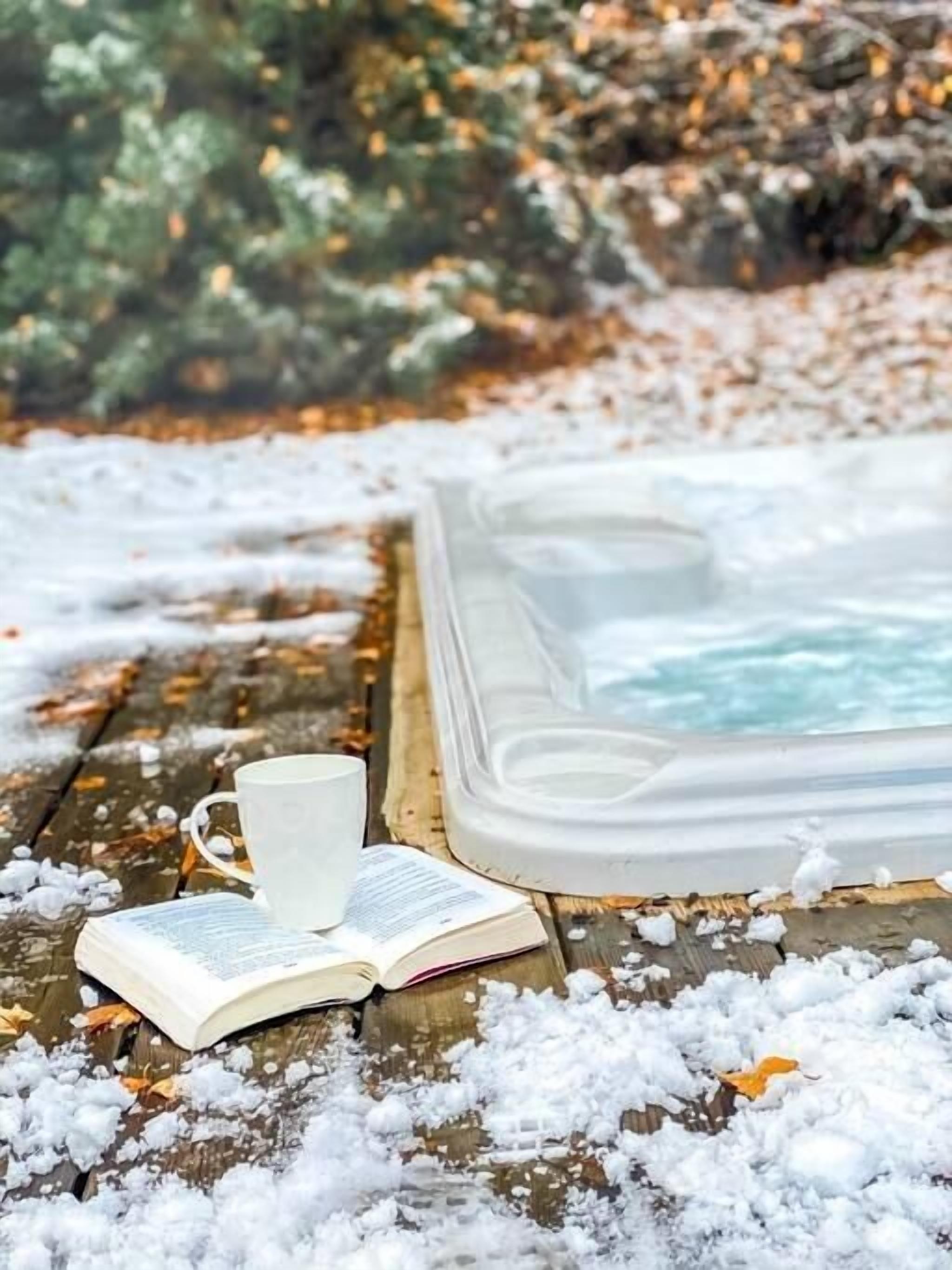Ein Whirlpool, umgeben von Schnee, ein Buch und eine Tasse auf einer Holzfläche.
