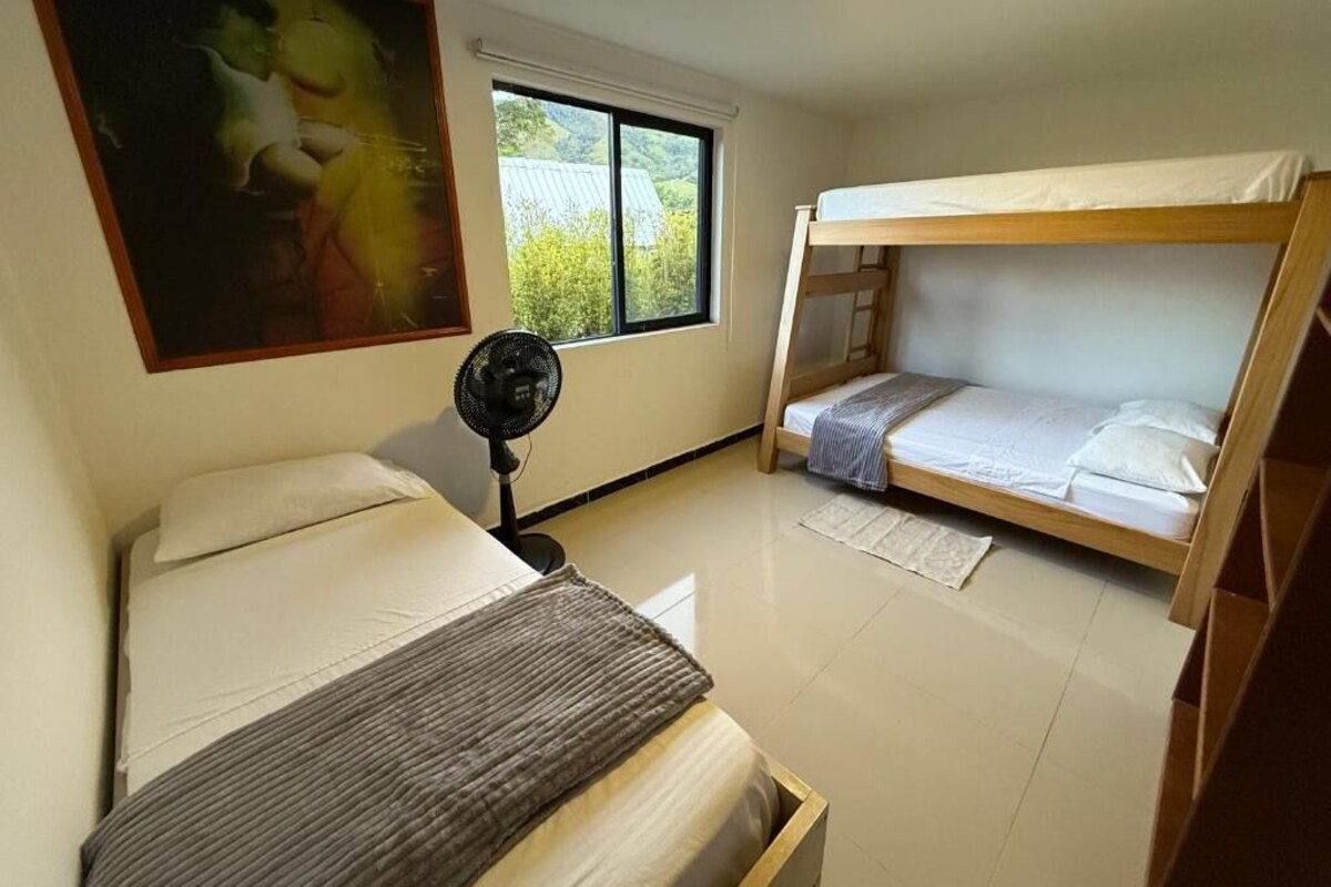 Una habitación con literas, una cama individual, un ventilador y una ventana con vistas a la vegetación.