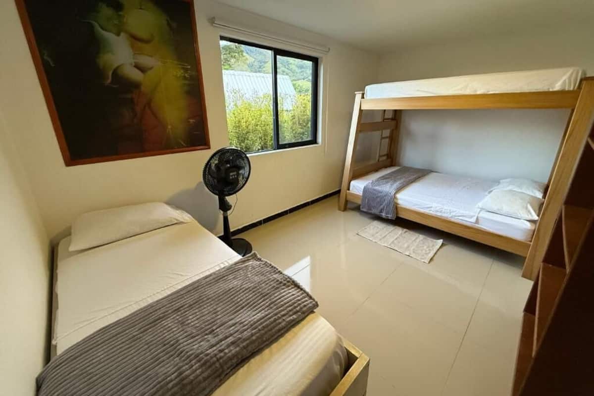 Une chambre avec un lit superposĂ©, un lit simple, un ventilateur et une fenĂȘtre donnant sur un paysage verdoyant.