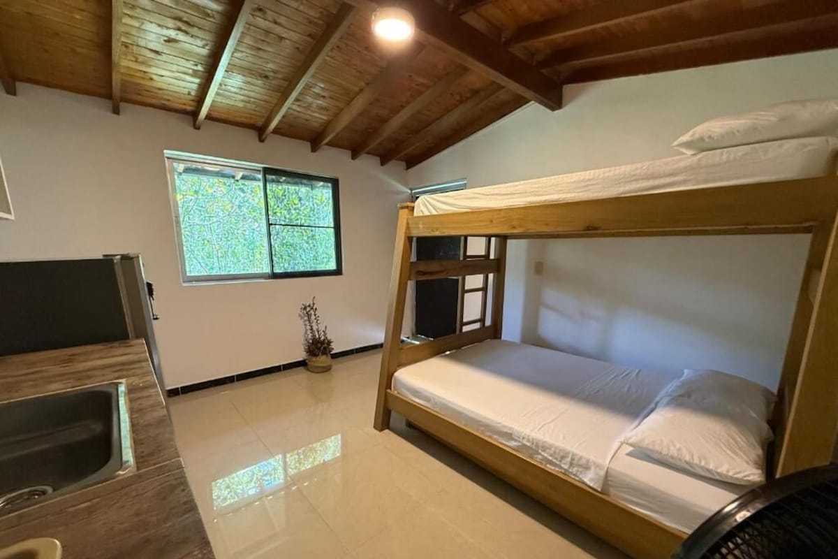 Dormitorio con literas, techo de madera, ventana, lavamanos y ventilador.