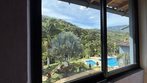 A window with a view of a landscaped garden, a swimming pool, and a building in the background. - Hotel Selva Cristal (San Carlos)