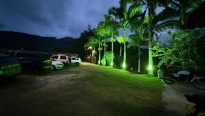 A parking area with several cars and motorcycles, surrounded by palm trees and a building with illuminated green lights. - Hotel Selva Cristal (San Carlos)