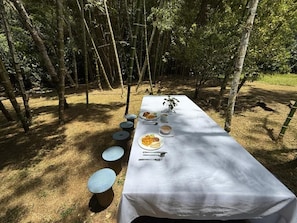 An outdoor dining table set up in a forested area with a white tablecloth, surrounded by chairs and plates of food.