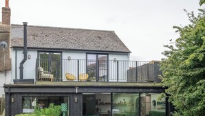 A modern house with a flat roof, a balcony with yellow chairs, and a glass door leading to a living area with a sofa and chairs. - Chic Devon Designer Oasis with hot tub (Kingsbridge)