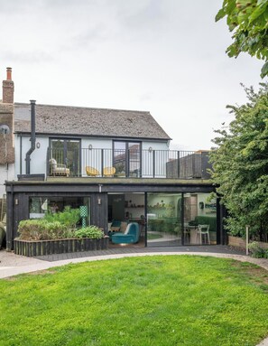 A modern house with a flat roof, a balcony with yellow chairs, and a glass door leading to a living area with a sofa and chairs. - Chic Devon Designer Oasis with hot tub (Kingsbridge)