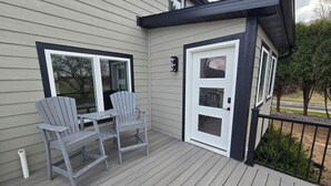 Una terraza de madera con dos sillas Adirondack y una pequeña mesa, una puerta con ventanal, y una barandilla de metal.
