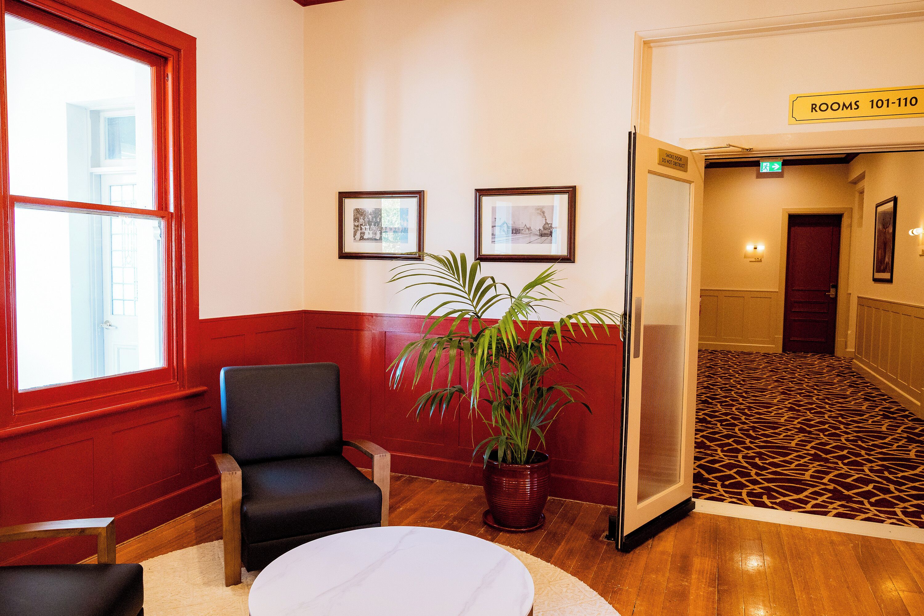 Un hall d’hôtel aux couleurs rouge et blanc, agrémenté d’une plante en pot, de tableaux encadrés accrochés au mur et d’un sol recouvert de moquette, qui m ène à une porte indiquant les numéros de chambre « 101 à 110 ».