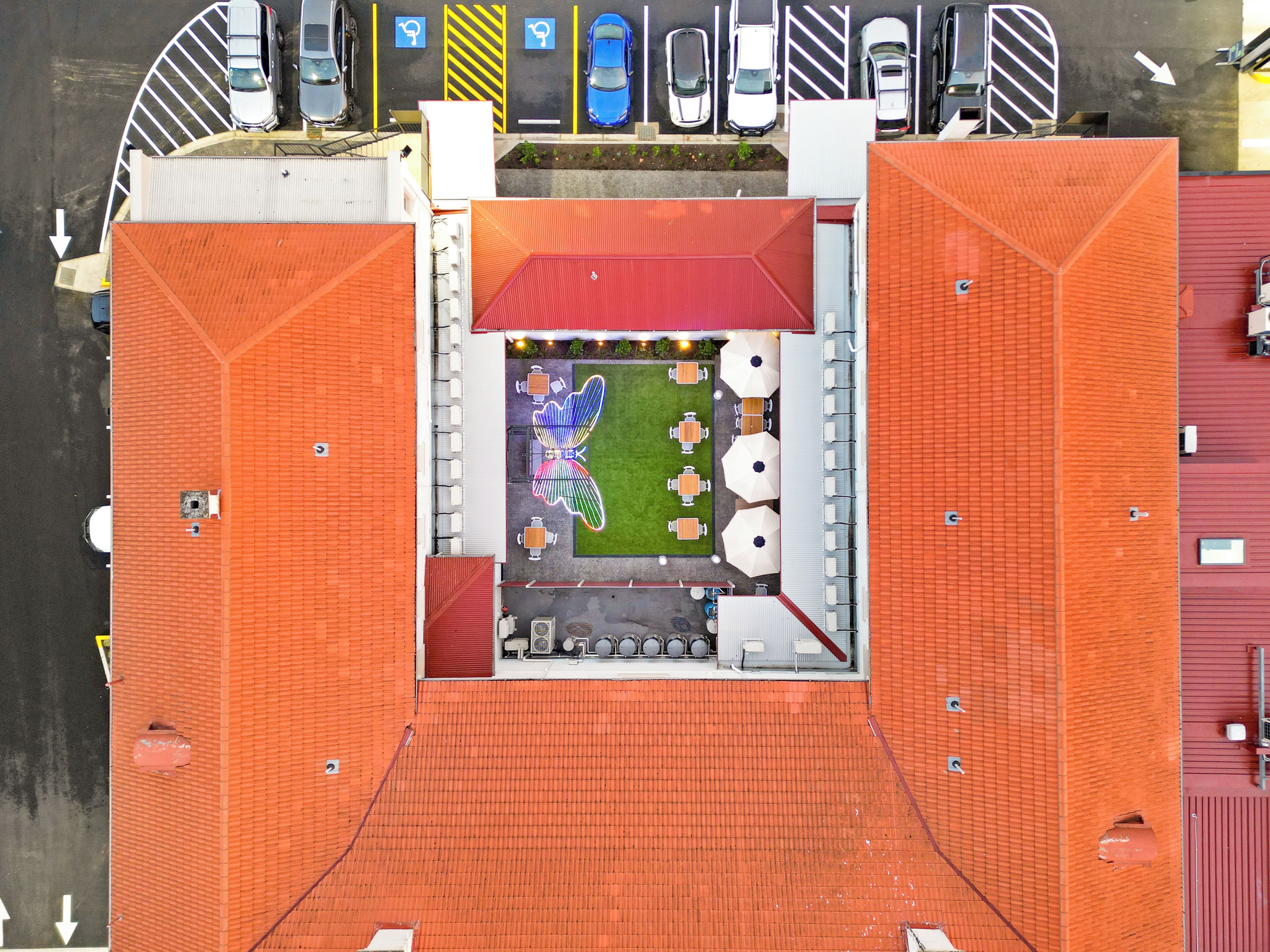 An aerial view of a parking lot with cars, a building with orange roofing, and a small outdoor seating area with tables and umbrellas.