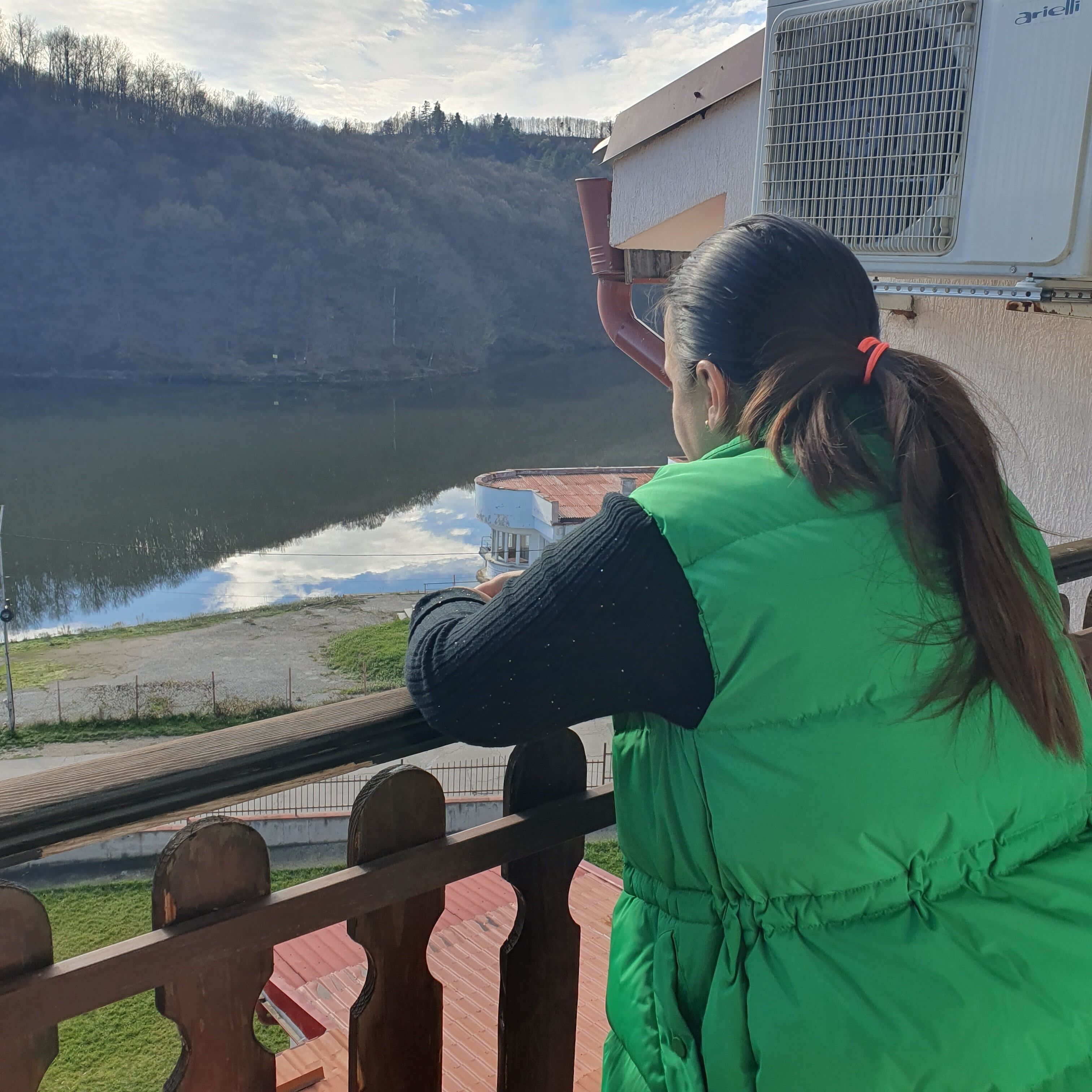 A person in a green vest standing by a railing overlooking a body of water with a hilly landscape in the background.