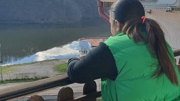 A person in a green vest standing by a railing overlooking a body of water with a hilly landscape in the background.