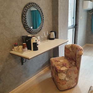 Una habitación con un escritorio largo y estrecho, un sillón floral, un espejo con marco decorativo, un hervidor, una cafetera y una ventana con vistas.