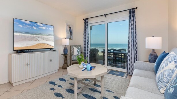 Ein Wohnzimmer mit Fernseher, einer Couch, einem Tisch mit Dekoration, Meerblick und einem Balkon mit Vorhängen.