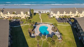 Luftaufnahme eines Resorts mit einem Swimmingpool, umgeben von Liegestühlen und Palmen, mit Blick auf einen Strand.