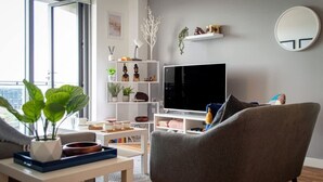 A modern living room with a grey armchair, a white TV stand, and a bookshelf with decorative items. - Modern 2BR Apartment w/ Balcony | Sleeps 6 (Merseyside)