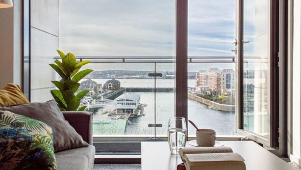A modern room with a large window overlooking a river and cityscape, a sofa with cushions, a glass of water, and a book on a table. - Modern 2BR Apartment w/ Balcony | Sleeps 6 (Merseyside)