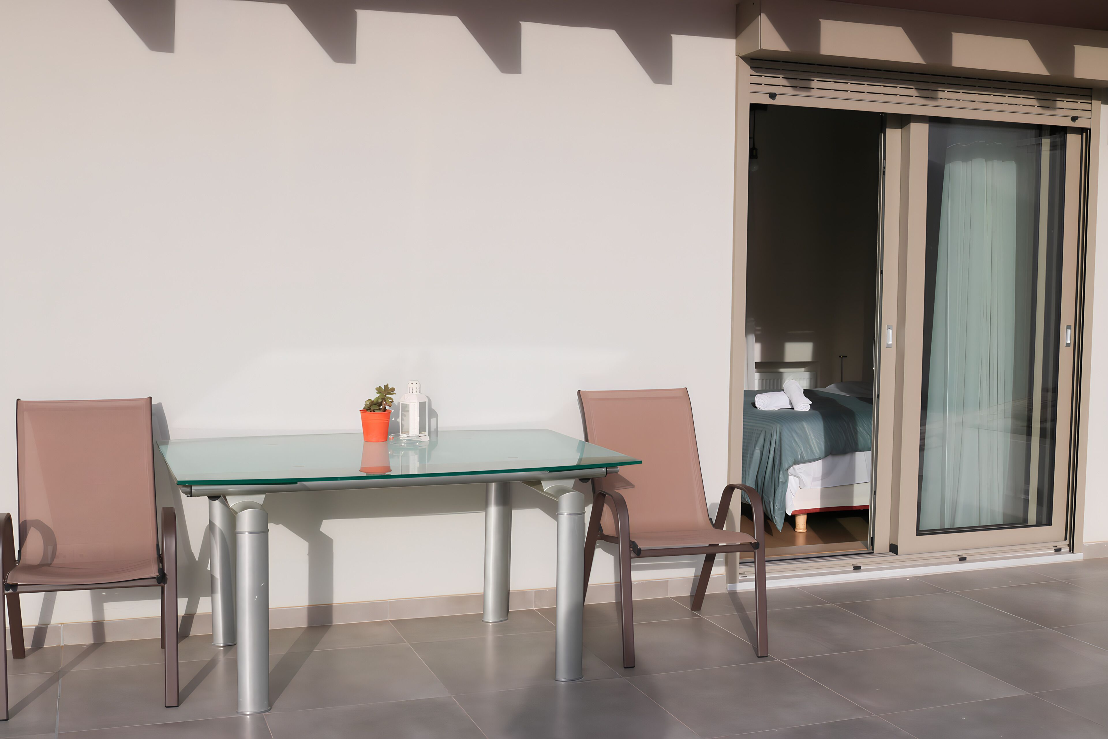 A glass-top table with two chairs, a potted plant, and a sliding glass door leading to a bedroom.
