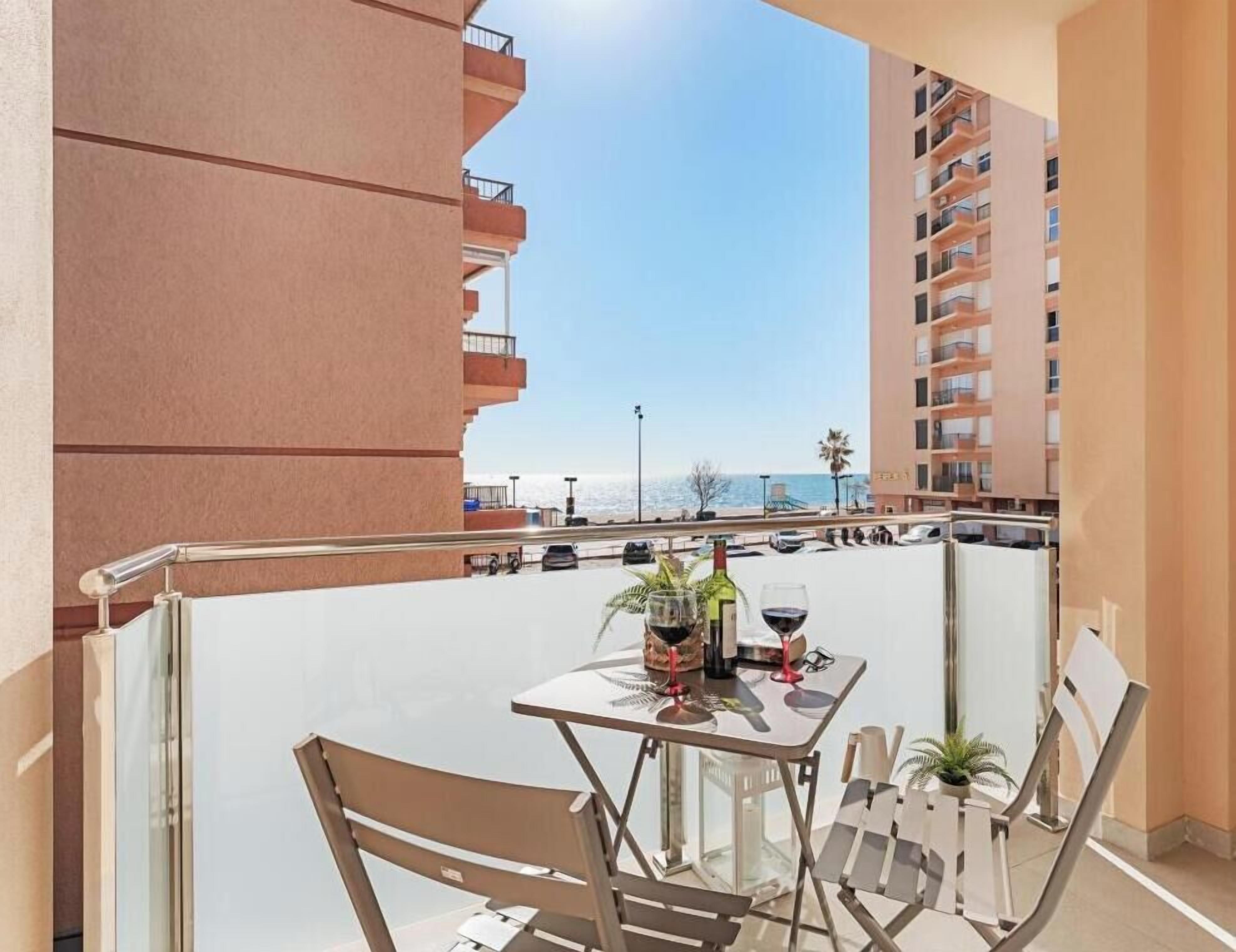 A balcony with a table set for two, outdoor seating, and a view of the sea and other buildings.