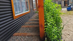 A building with a corrugated exterior, wooden steps leading to a door, and a small garden area with green plants. - Apart Hotel Plaza Araucaria, Individual Apartment 5 p. Mañihuales, Carretera Austral (Villa Mañihuales)