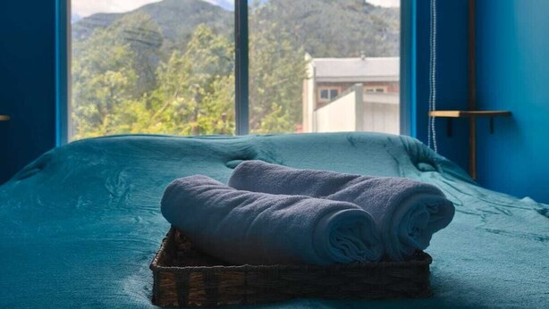 A room with a bed, a window with a view of mountains, and a basket with rolled towels. - Apart Hotel Plaza Araucaria, Individual Apartment 5 p. Mañihuales, Carretera Austral (Villa Mañihuales)