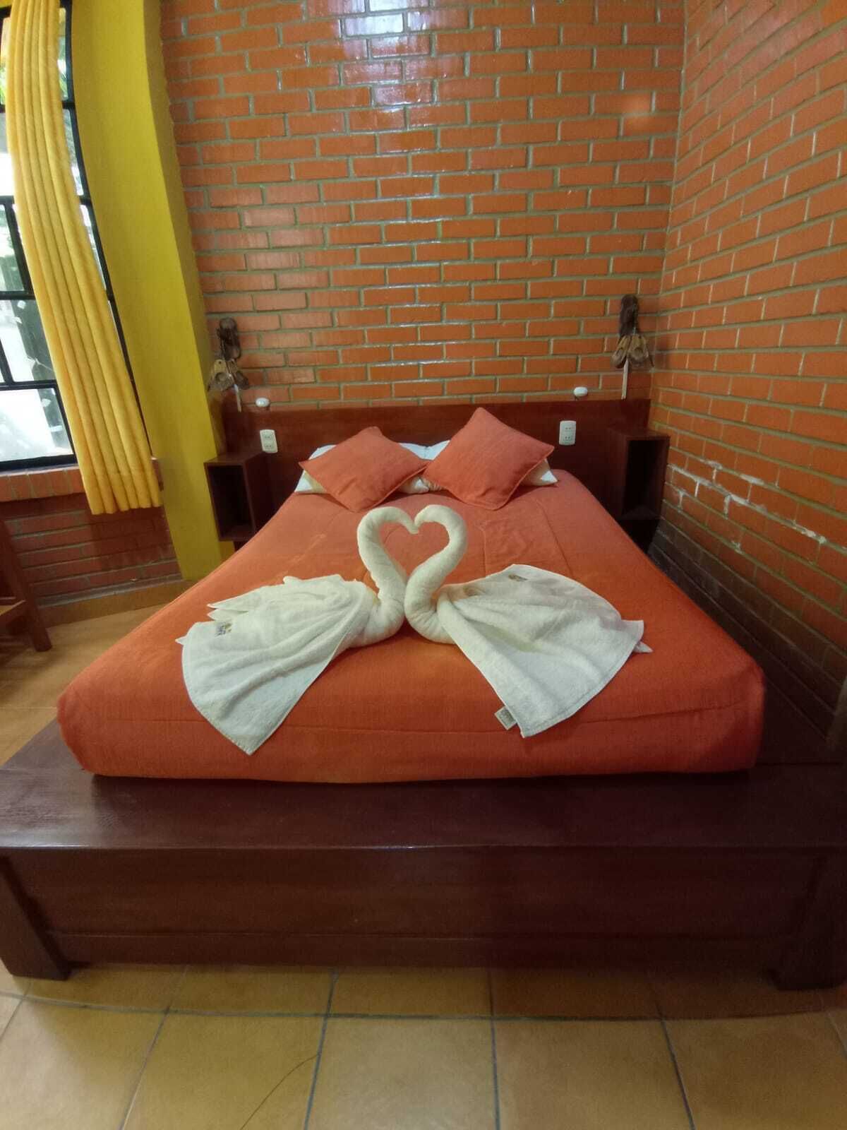 Una cama con una toalla dispuesta en forma de corazón, un cabecero de madera y una pared de ladrillo.