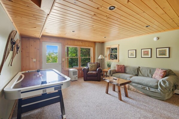A cozy living room with a pool table, a sofa, and a chair. There are windows with a view of greenery outside.
