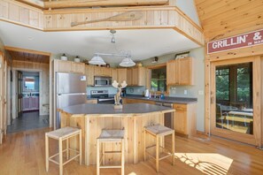 A cabin-style kitchen with a central island, wooden bar stools, and a sign for 'Grillin' & In Maine'. - Lake View House with Hot Tub, Sauna and Game Loft (Woodstock)
