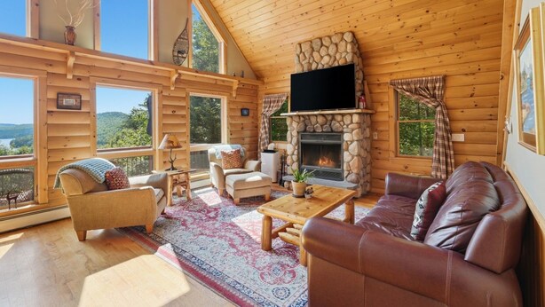 Interior de una cabaña de troncos con chimenea de piedra, mobiliario de madera y ventanas amplias que ofrecen vistas al exterior.