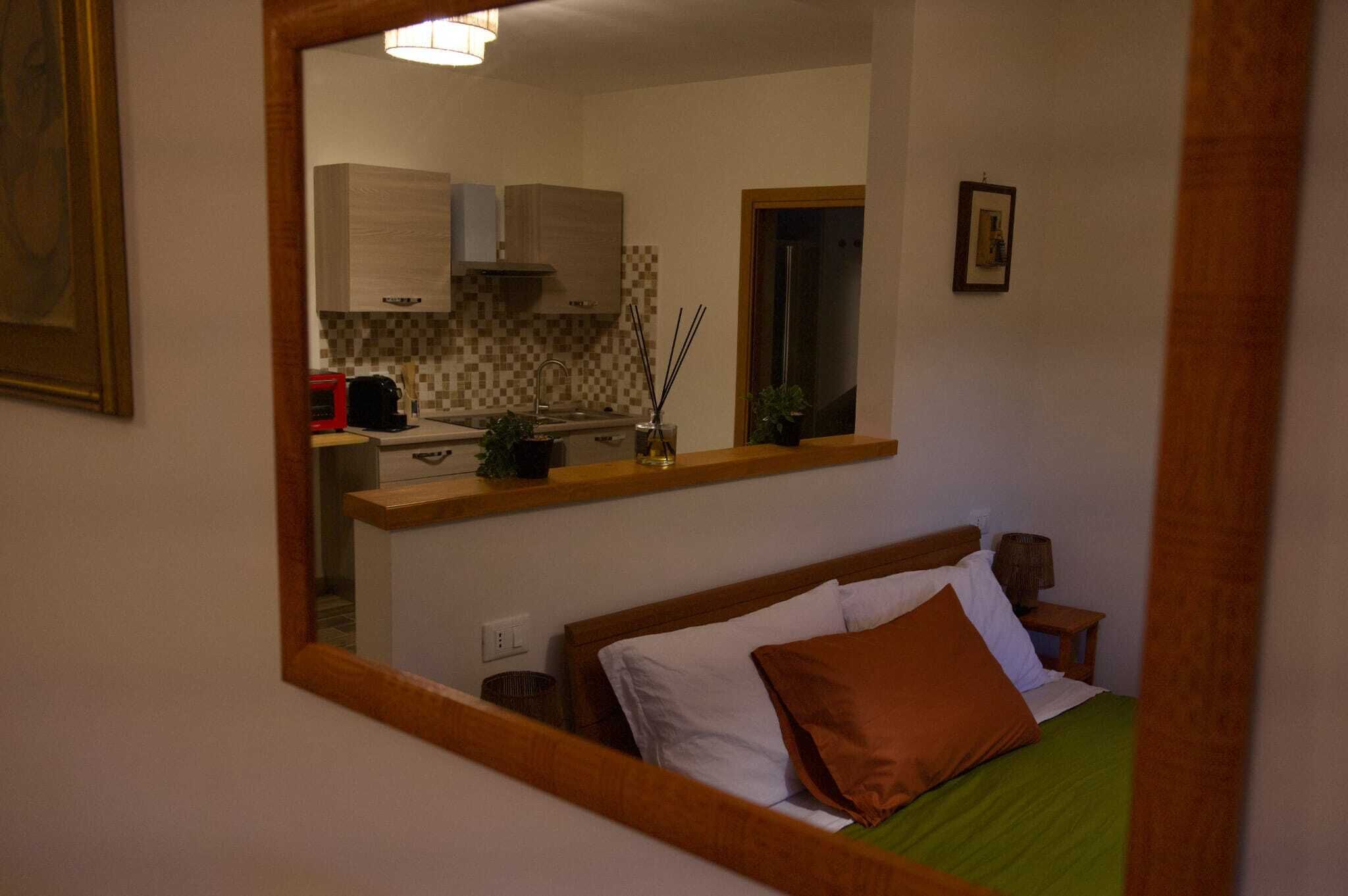 A bedroom with a bed, a nightstand, a mirror reflecting a kitchen area with cabinets and a sink.
