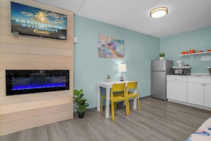 A room with a TV displaying 'Latitude 26° Waterfront Resorts,' a fireplace, a dining table with yellow chairs, a small plant, and a kitchen area with a refrigerator and microwave. - 2 Queen with Kitchenette Garden view in Fort Myers Beach (Fort Myers Beach)