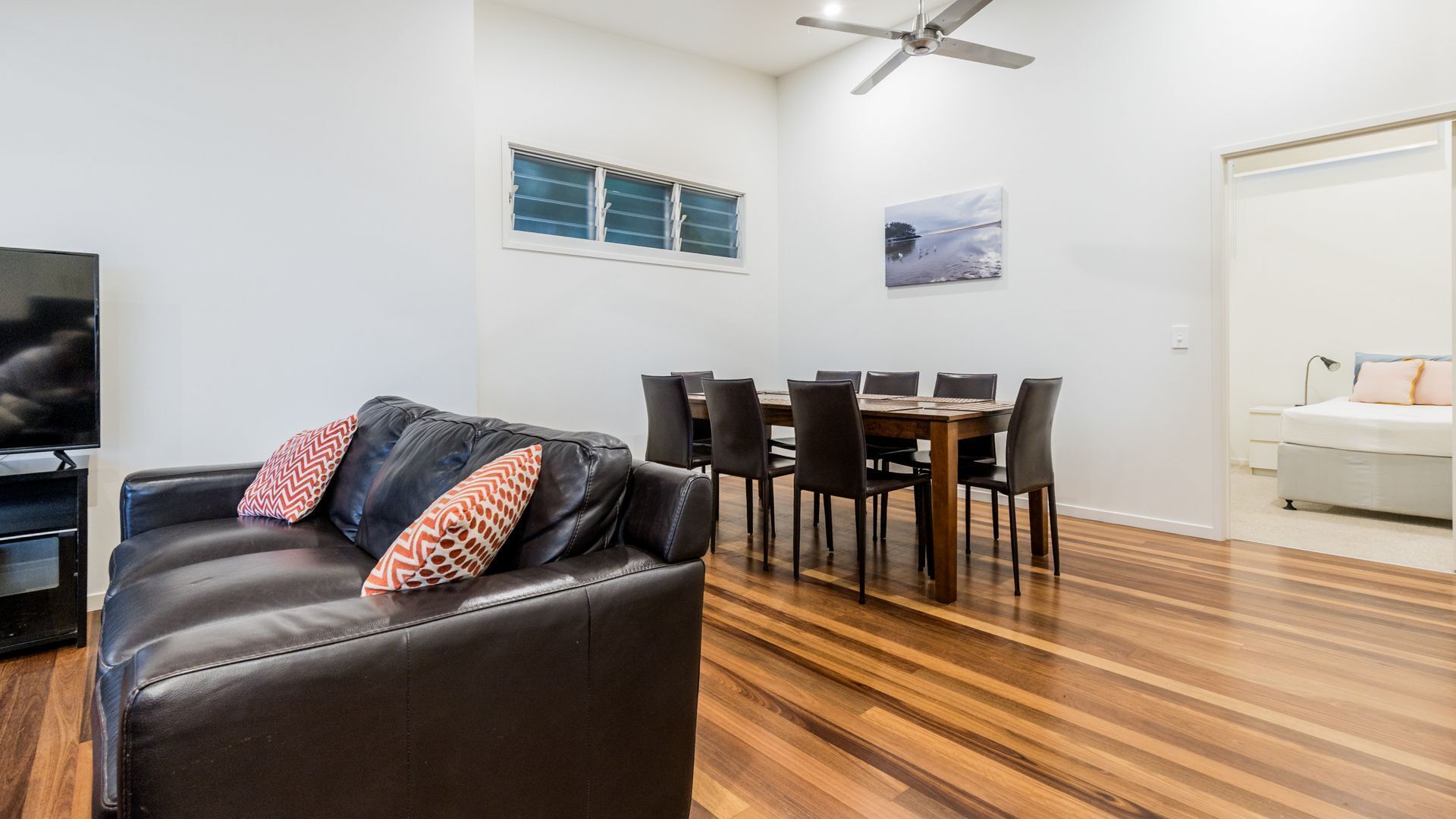 Uma sala de estar moderna com sofá de couro, mesa de jantar, cadeiras, ventilador de teto, televisão e uma cama.