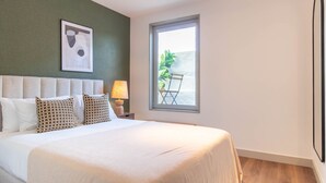 Ett hotellrum med en säng, ett nattduksbord med en lampa, ett fönster med utsikt över grönområden och en inramad tavla på väggen.