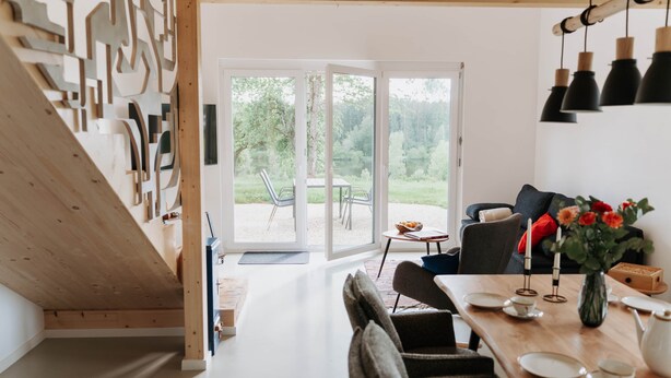 A modern interior with a wooden staircase, a dining area with a table set for two, and a view of the outdoors through glass doors.
