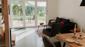 A modern living room with a sliding glass door leading to an outdoor patio, a dark-colored sofa, a wooden dining table with a candle, and a small table with a bowl and a chair.
