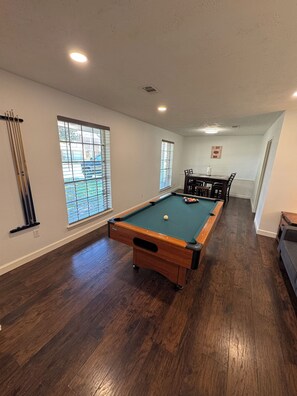 Una mesa de billar en una sala con pisos de madera, ventanas amplias y una zona de comedor.