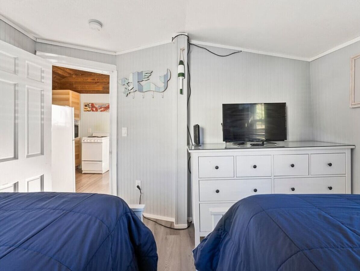 Une chambre avec deux lits, un téléviseur posé sur une commode, et une cuisine visible à travers une porte ouverte.