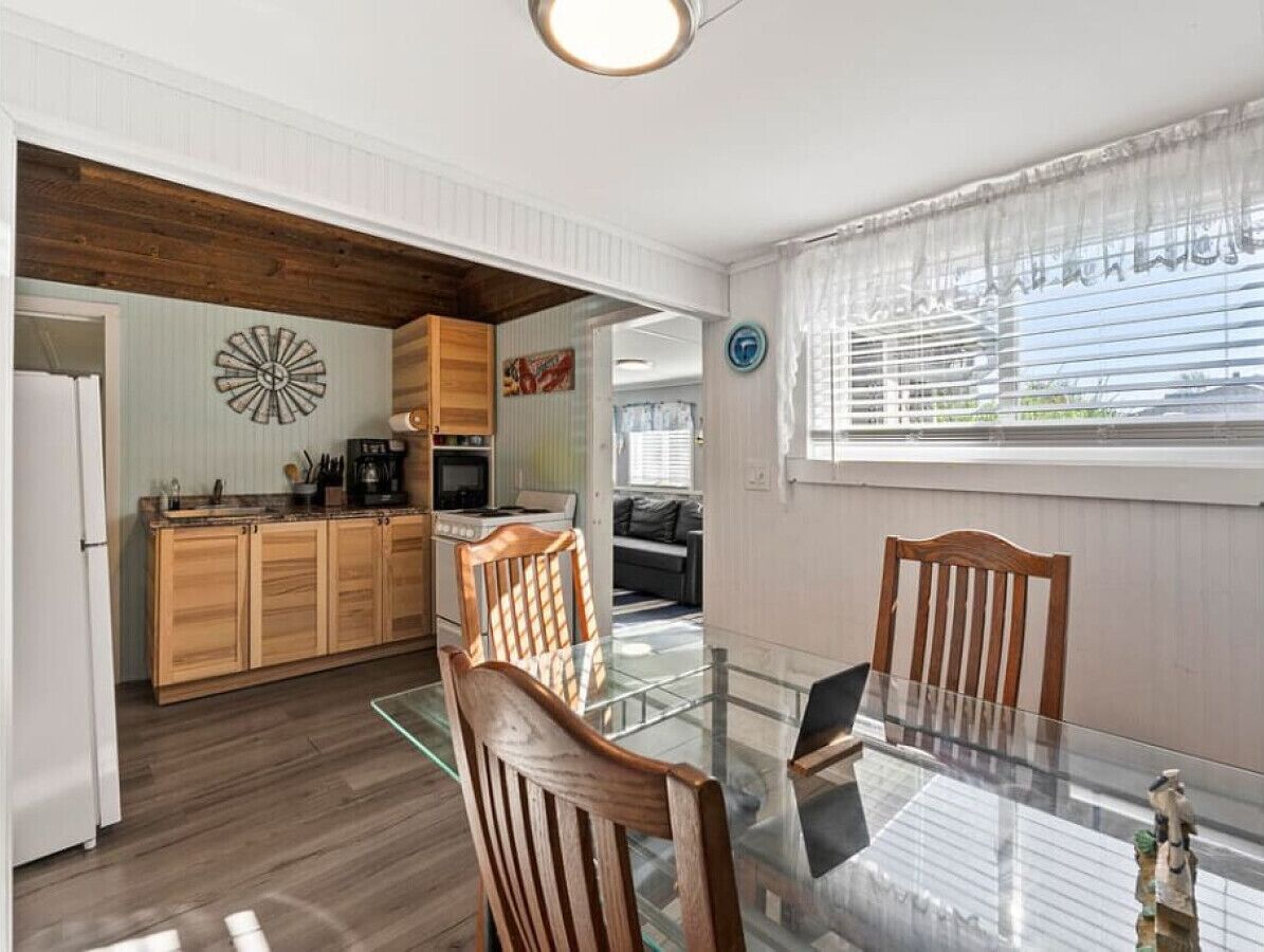 A compact kitchen with wooden cabinets, a dining table with chairs, a coffee maker, and a clock on the wall.