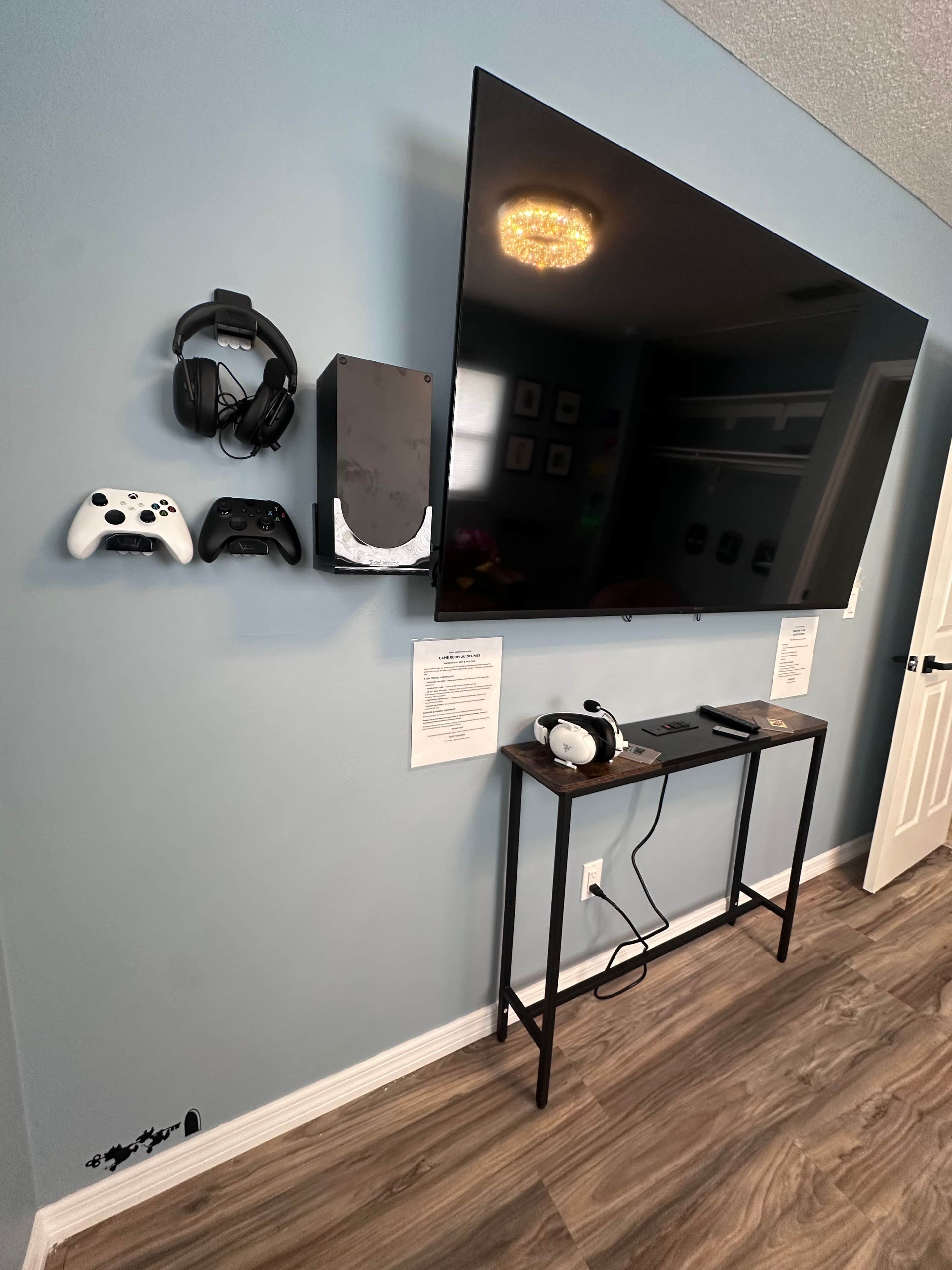 Een tv aan de muur, met gameconsoles en hoofdtelefoons, een klein tafeltje met een headset, en een deur met een bordje.