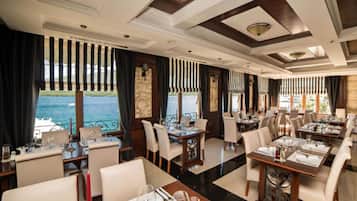 Un restaurant où les tables sont dressées pour le service, de grandes fenêtres offrant une vue sur la mer et un design moderne au niveau du plafond.