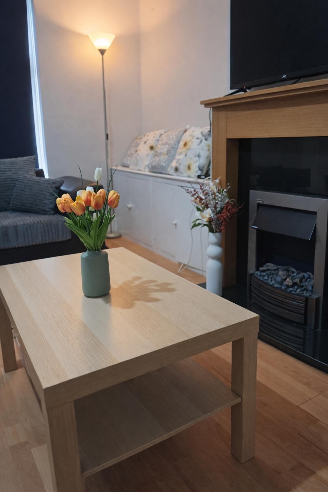 A modern living room with a wooden table, a vase of flowers, a sofa, a fireplace, and a lamp.