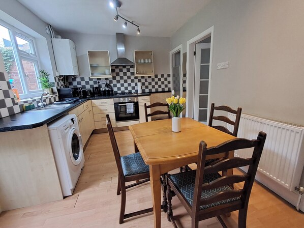 A compact kitchen with a dining table, chairs, a washing machine, and a radiator. - 2 Bed Home - Parking, Wifi, Smart Tv (Nottingham)