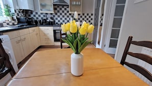 A kitchen with a checkered backsplash, a white vase with yellow flowers on the table, and a wooden chair. - 2 Bed Home - Parking, Wifi, Smart Tv (Nottingham)