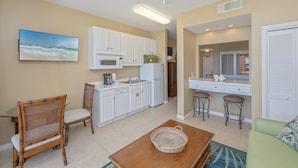 A compact kitchen with white cabinets, a microwave, and a refrigerator. There is a dining area with wicker chairs and a wooden table. A television is mounted on the wall, and a ceiling fan is visible. - Destin West V302 (Fort Walton Beach)