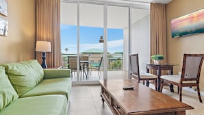 A hotel room with a green sofa, a wooden coffee table, a TV mounted on the wall, a balcony with a table and chairs, and a view of the sky. - Destin West V302 (Fort Walton Beach)