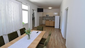 A dining area with a long wooden table, chairs, and a vase with flowers. A kitchen is visible in the background with cabinets and a refrigerator. - Sunset Bay Villa Kevin (Orebic)
