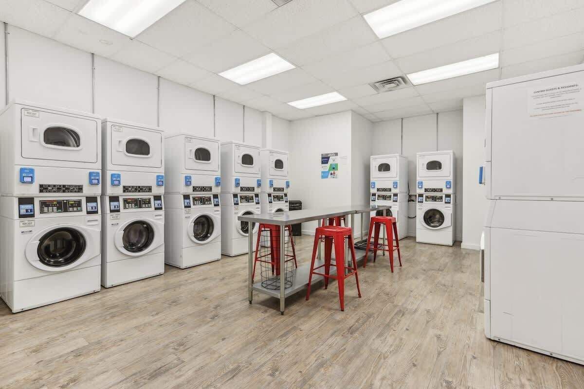 A laundry facility with multiple washing machines, a central table with stools, and a whiteboard on the wall.