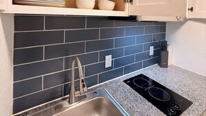 Een keuken met een RVS spoelbak, een zwart inductiekookplaatje, een donkere betegelde spatwand en houten planken met servies en glazen.