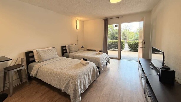 A room with two beds, a desk, a chair, a TV, and a sliding glass door leading to an outdoor area. - [27] ATX Studio: 2 Twin Beds, Pool, Gym & Dog Park (Austin)
