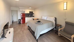 Ein kleines Zimmer mit Bett, Tisch, einer Sitzecke, einer Küchenzeile und einem Fernseher.